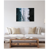 Framed aerial photo of ocean waves crashing on Iceland's black beach, showcased above a beige sofa with striped and solid cushions in a neutral living room.