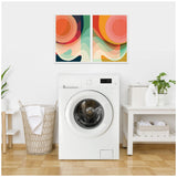 Two framed abstract canvas paintings with overlapping circular shapes in warm orange, pink, green, and beige hues displayed side by side on a white wall above a white front-load washing machine in a laundry room.