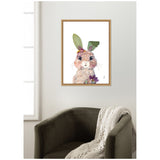 Framed watercolor print of a bunny with large eyes, leafy ears, and small flowers around its head, hanging above a dark olive green armchair draped with a cream knit blanket.