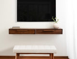 Wall-mounted floating console table with two drawers in warm wood tones, topped with a small plant and books beneath a large flat-screen TV.