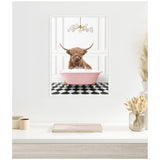 Framed canvas showing a brown Highland cow sitting in a pink clawfoot bathtub filled with bubbles on a black-and-white checker floor under a modern gold chandelier