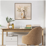 A beige fabric chair faces a wooden desk with books, storage boxes, and vases, beneath a framed canvas showing a rustic pitcher holding white and beige flowers with green leaves.