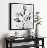 Black and white framed canvas of a large succulent close-up above a black console table with stacked books, a geometric star sculpture, a round clock, and a patterned white vase with green branches.
