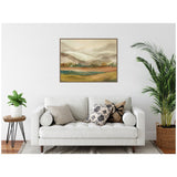 Muted earth-toned mountain landscape painting with soft green and brown hues, displayed above a white sofa accented by patterned pillows and indoor plants.