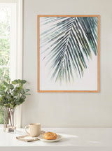 Framed close-up print of a green palm leaf with white background, hanging on a light gray wall above a table with a plant, coffee cup, and croissant.
