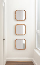 Three square wooden-framed mirrors with rounded corners arranged vertically on a white wall beside a window, reflecting parts of the room and window light.