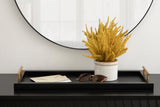 Black rectangular tray with brass handles holding a white planter with yellow dried flowers, sunglasses, and two beige envelopes on a black surface beneath a round mirror.