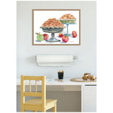 Framed watercolor painting displaying two golden brown apple pies on gray pedestal stands with red and green apples scattered around, hanging above a light-colored desk and chair.