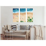 Three-panel wall art showing a beach scene with sandy shore, turquoise water, surfboards, and palm trees, displayed above a beige couch with patterned pillows.