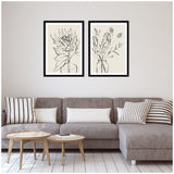 Two black-framed minimalist ink line drawings of protea flowers in vases, hung side by side above a gray sectional sofa with striped and solid pillows.