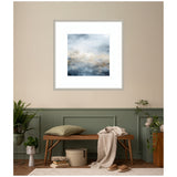 Abstract wall art print with soft blue, beige, and gray tones depicting a serene oceanic scene, framed in white, hanging above a wooden bench with a pillow and draped throw.