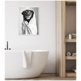 Black and white framed canvas of a lion wrapped in a towel, hanging above a white freestanding bathtub with a towel draped over its edge in a minimalist bathroom.