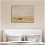 Framed canvas print of a soft pastel landscape showing a desert scene with sandy tones, muted green shrubs, and a pale pink sky, hung above a bed with neutral beige cushions.