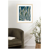 Wall art of elongated beige and green leaves on dark blue background, framed in light wood, hanging above a dark olive green armchair with an open book and next to a potted leafy plant.