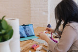 Person arranging colorful yarn skeins on a wooden table with a tablet displaying instructions nearby, in a softly lit room with brick wall.