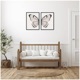 Two-piece framed canvas art showing symmetrical black and white watercolor butterfly wings on a neutral linen background, above a wooden bench with white cushions
