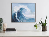 Framed abstract painting of a large blue ocean wave cresting with white foam, displayed above a white table with plants and books.