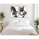 Two framed black and white abstract art pieces with bold, intertwined brushstrokes hang above a white bed in a minimalist bedroom with leafy green plants.