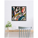 Square wall art with abstract, colorful ficus leaves in shades of teal, olive, mustard, coral, and pink framed in black above a light wood desk with a plant and gray chair.