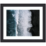 Aerial view of dark ocean waves crashing onto a black sand beach, with white foam creating intricate patterns between water and shore.