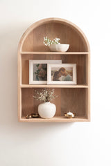 Light wood wall shelf with a rounded arch top, holding white bowls with flowers, two framed photos, a white vase with small flowers, and small brass objects.