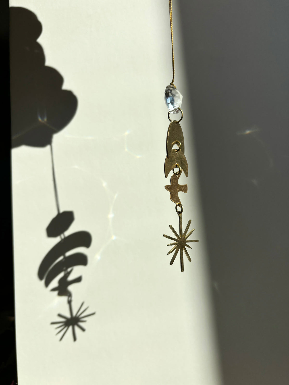 Hanging ornament featuring a clear vintage glass crystal atop three gold geometric shapes, casting a distinct shadow on a light background.