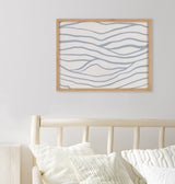 Framed art print with soft blue wavy lines mimicking coastal waves, hanging above a light wood slatted bench with textured cream pillows.
