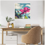 Framed canvas of a vivid pink clematis flower and buds against a partly cloudy blue sky, hanging above a wooden desk with a beige chair and minimal decor.