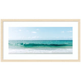 Framed panoramic photo of a turquoise ocean wave breaking under a clear blue sky with scattered clouds, bordered by white sandy shore visible at the bottom.