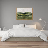 Abstract green mountain landscape painting with soft brushstrokes in a light wooden frame over a white bed with pillows and a modern adjustable lamp to the right.