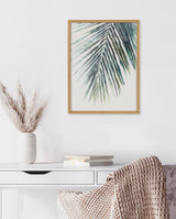 Framed botanical print of green palm leaves with blue accents on a white wall above a white desk with stacked books and a textured beige throw on a chair.