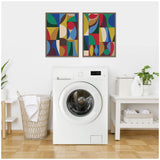 Two framed abstract paintings with colorful geometric shapes in blue, red, green, yellow, and brown above a white front-loading washing machine in a laundry room.