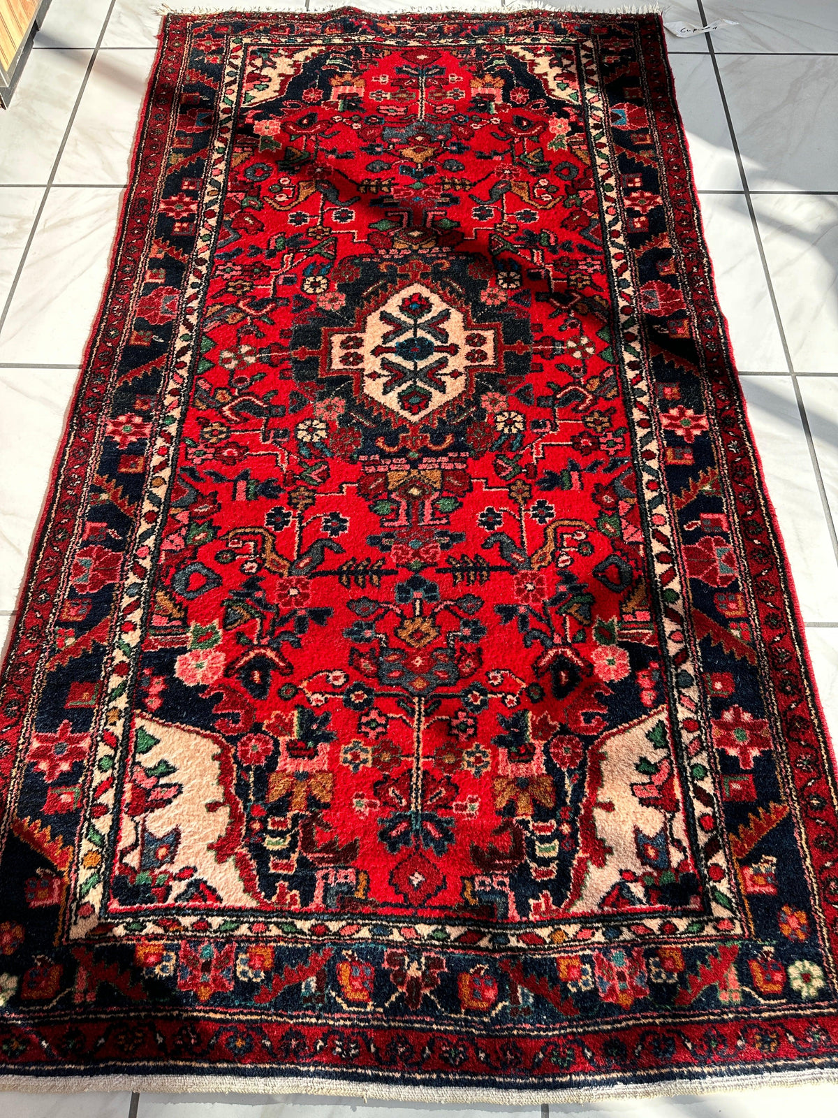 Rectangular hand-knotted Persian Hamadan wool rug featuring a red field with intricate floral motifs and a central cream medallion, bordered by navy blue and cream patterns.