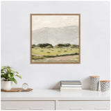 Framed square canvas print showing a muted landscape with green fields, small dark bushes, and soft gray-blue mountains under a pale cloudy sky, hanging above a white dresser with books, plant, and patterned cups.
