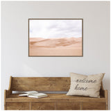 Framed canvas print of soft pink desert sand dunes under a cloudy sky, hung above a rustic wooden bench with a "welcome home" pillow and open book.