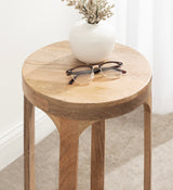 Round wooden drink table with smooth grain and natural finish, holding a white ceramic vase with dried flowers and a pair of tortoiseshell eyeglasses.