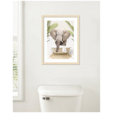 Framed print of an elephant facing forward in a white vintage bathtub with palm leaves framing the scene, hung above a white toilet against a pale wall.