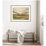 Softly colored horizontal painting of a misty mountain landscape with green, brown, and beige tones, framed in light wood and hung above a bench with a book and draped fabric.
