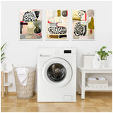 Three framed abstract canvases with organic shapes in beige, black, white, red, and olive tones hung above a white front-loading washing machine in a minimalist laundry room.