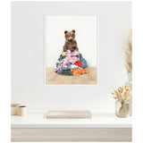 Framed canvas artwork of a small brown bear sitting atop a colorful pile of laundry on a light wooden floor against a white wall, displayed in a minimalist room with dried plants.