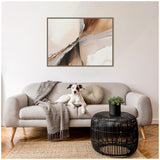 Light gray sofa with wooden legs, adorned by a hound dog, a round cushion, and a fringed throw; a black round woven coffee table with a small green plant on top rests on a natural fiber rug