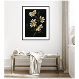 Framed botanical artwork of cream-colored flowers with green stems on a dark background, hanging above a wooden bench with a draped light blanket and an open book.