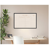 Black-framed wall pinboard resembling vintage linen postcard design, hanging above light wood desk with white chair, small plant, and stacked books.