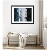 Framed aerial photo of ocean waves crashing onto a black sand beach in Iceland, hanging on a white wall above a wooden bench with an open book and draped fabric.
