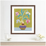 Colorful framed art print of snowdrop flowers with blue, pink, and white petals in a patterned vase against an olive green and light blue background, hanging above a white shelf.
