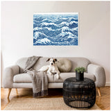 Blue and white abstract linocut print of ocean waves in a white frame, hung above a light gray sofa with a textured gray throw and a round beige pillow