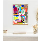 Abstract painting with colorful geometric and organic shapes including red, blue, green, mustard, and black, framed in light wood and hung on a white wall above a minimalist desk.