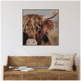 Close-up of a highland cow with shaggy reddish-brown fur and a light brown horn, set in a simple wooden frame above a rustic bench with a 'welcome home' pillow.