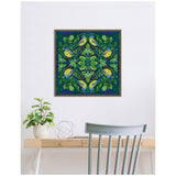 Square framed canvas art featuring symmetrical pattern of four yellow and blue birds surrounded by green leaves and flowers on a deep blue background, hanging above a wooden desk with a plant.