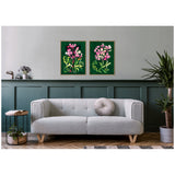Two framed canvases with pink and yellow abstract flowers on rich green backgrounds hanging above a light gray sofa with a round gray pillow in a minimalist living room featuring paneled green walls and wood flooring.